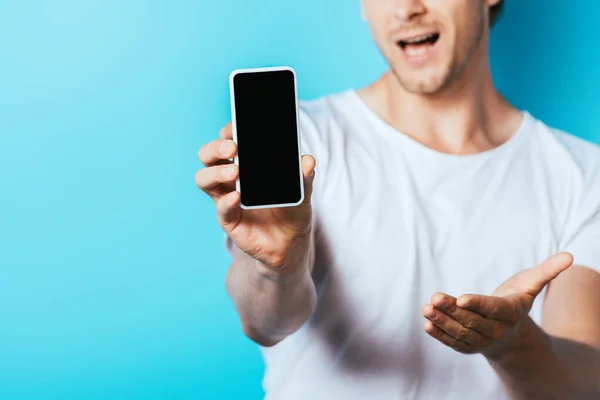 Vista recortada del hombre apuntando con la mano mientras muestra el teléfono inteligente con pantalla en blanco sobre fondo azul - foto de stock