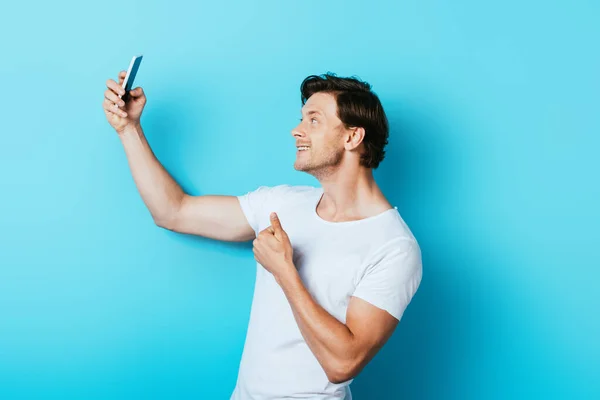 Young man in white t-shirt showing thumb up while taking selfie with smartphone on blue background — Stock Photo