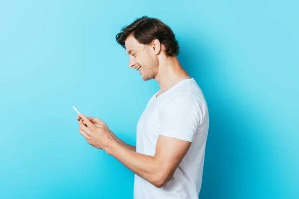 Vista lateral del joven en camiseta blanca con smartphone sobre fondo azul - foto de stock