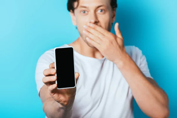 Selective focus of shocked man showing smartphone with blank screen on blue background — Stock Photo