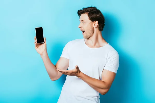 Homem surpreso apontando com a mão enquanto segurava o smartphone com tela em branco no fundo azul — Fotografia de Stock