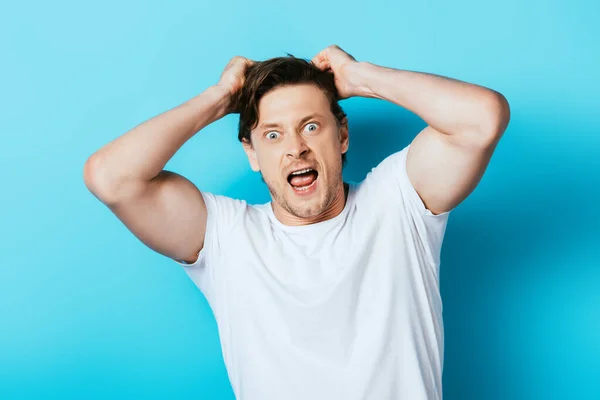 Homme fou avec les mains près des cheveux en t-shirt blanc regardant la caméra sur fond bleu — Photo de stock