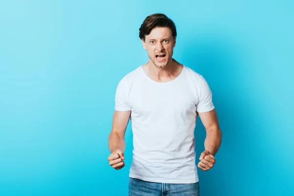 Irrated man in white t-shirt with hands in fists looking at camera on blue background — Stock Photo