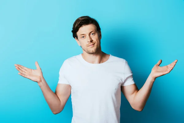 Hombre en camiseta blanca mostrando gesto de encogimiento sobre fondo azul - foto de stock