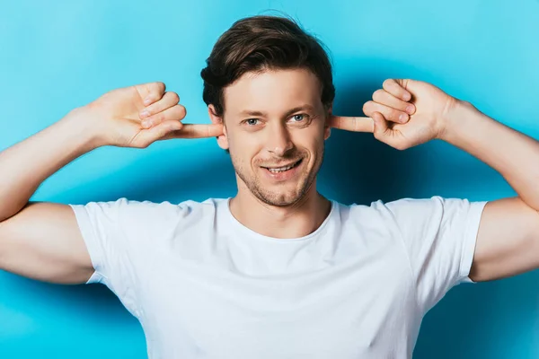 Hombre joven en camiseta blanca que cubre las orejas con los dedos sobre fondo azul - foto de stock