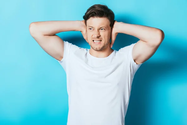 Homme en colère en t-shirt blanc couvrant les oreilles avec les mains sur fond bleu — Photo de stock