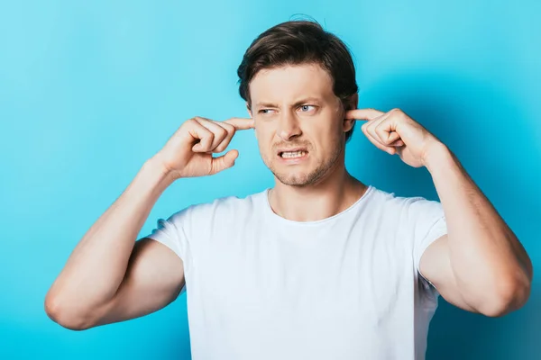 Homme agressif en t-shirt blanc couvrant les oreilles avec les doigts sur fond bleu — Photo de stock