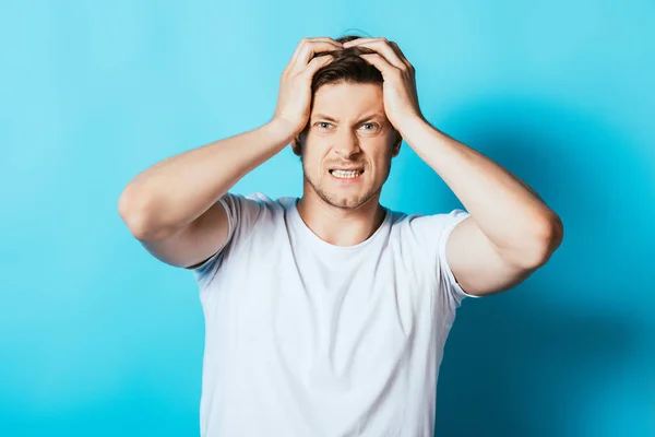 Joven hombre enojado en camiseta blanca mirando a la cámara sobre fondo azul - foto de stock