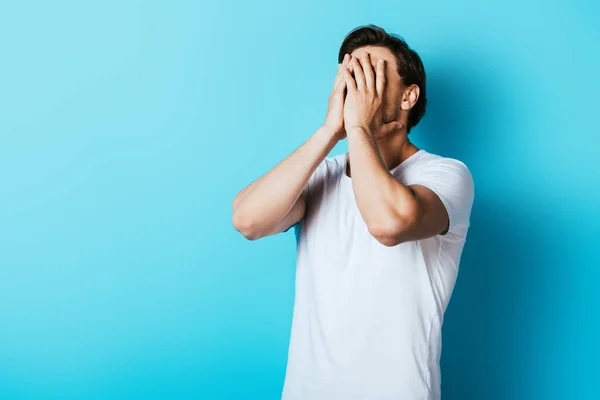 Scared man in white t-shirt covering face with hands on blue background — Stock Photo
