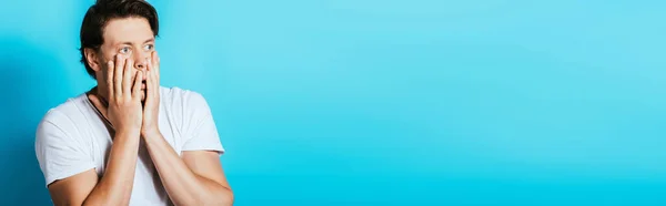Panoramic crop of surprised man with hands near cheeks on blue background — Stock Photo