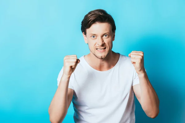 Wütender Mann im weißen T-Shirt mit Fäusten und Blick in die Kamera auf blauem Hintergrund — Stockfoto