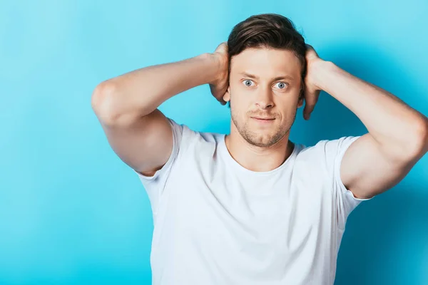 Hombre enojado en camiseta blanca cubriendo las orejas con las manos sobre fondo azul - foto de stock