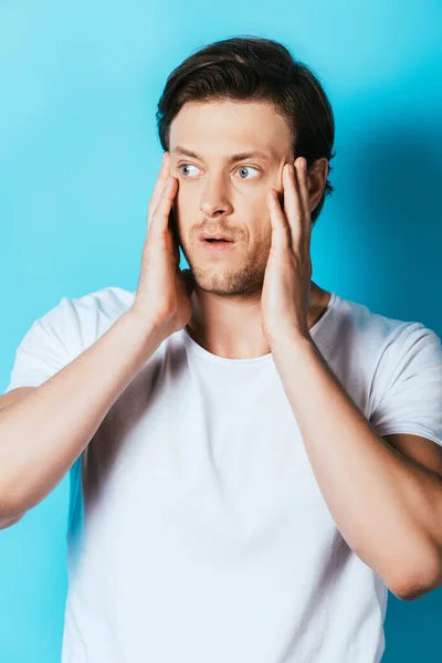 Hombre preocupado en camiseta blanca con las manos cerca de la cara sobre fondo azul - foto de stock