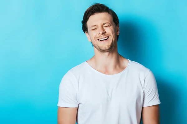 Young man in white t-shirt laughing with closed eyes on blue background — Stock Photo