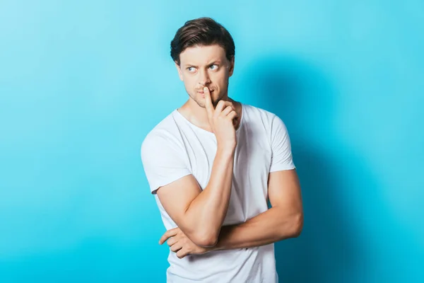 Hombre pensativo en camiseta blanca con el dedo cerca de los labios sobre fondo azul - foto de stock