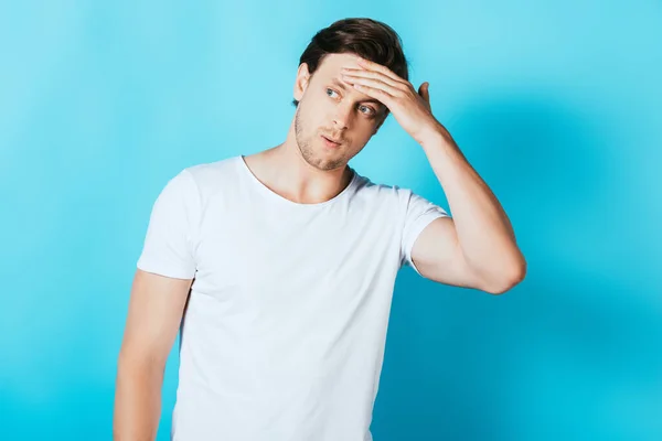 Giovane uomo in t-shirt bianca che tocca la fronte mentre distoglie lo sguardo su sfondo blu — Foto stock