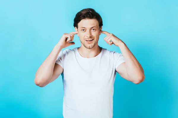 Junger Mann blickt in die Kamera, während er die Ohren mit Fingern auf blauem Hintergrund bedeckt — Stockfoto