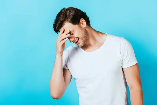 Junger Mann im weißen T-Shirt mit der Hand in Stirnnähe und Blick auf blauen Hintergrund — Stockfoto