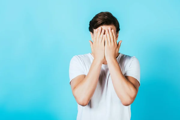 Frightened man in white t-shirt covering face with hands on blue background — Stock Photo