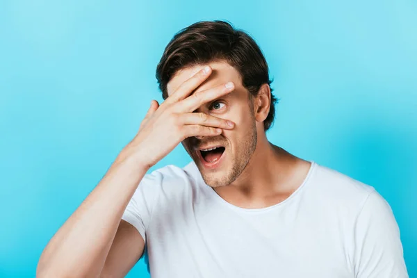 Young man looking away while covering face with hand isolated on blue — Stock Photo