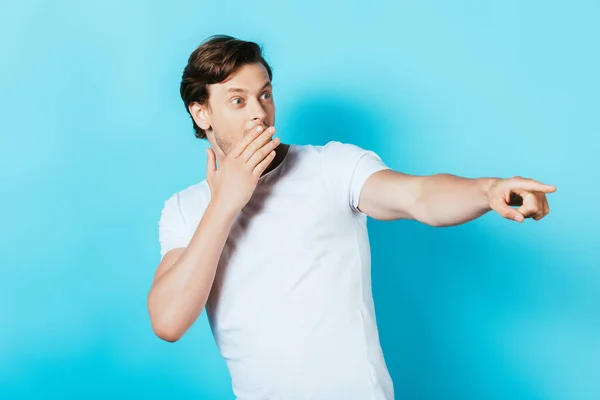 Hombre sorprendido cubriendo la boca con la mano y señalando con el dedo sobre fondo azul - foto de stock