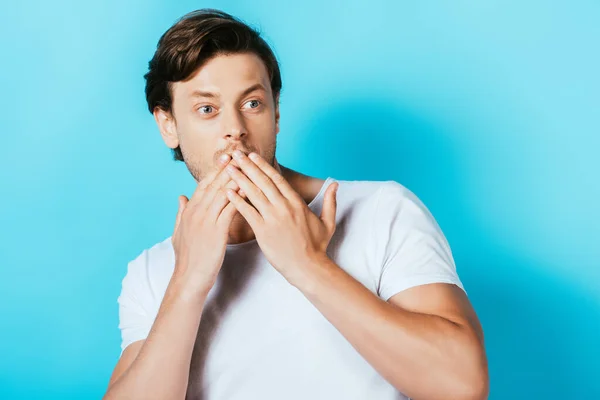 Homme choqué en t-shirt blanc couvrant la bouche avec les mains sur fond bleu — Photo de stock