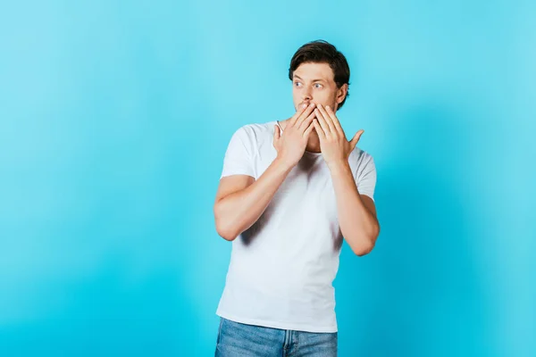Homem surpreso em branco t-shirt cobrindo boca com as mãos no fundo azul — Fotografia de Stock