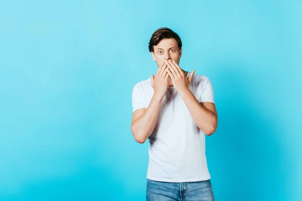 Schockierter junger Mann im weißen T-Shirt blickt in die Kamera auf blauem Hintergrund — Stockfoto