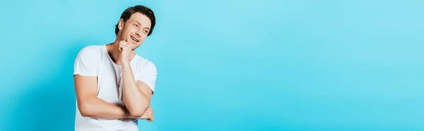 Panoramic crop of man in white t-shirt with fist near chin looking away on blue background — Stock Photo