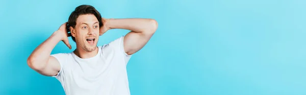 Panoramic shot of ecited man with hands near head looking away on blue background — Stock Photo