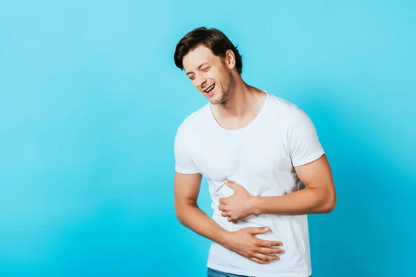 Hombre joven en camiseta blanca riendo y tocando el vientre sobre fondo azul - foto de stock