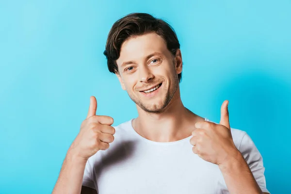 Hombre joven en camiseta blanca mostrando pulgares hacia arriba en la cámara sobre fondo azul - foto de stock