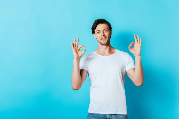Junger Mann im weißen T-Shirt zeigt Okay-Geste vor Kamera auf blauem Hintergrund — Stockfoto