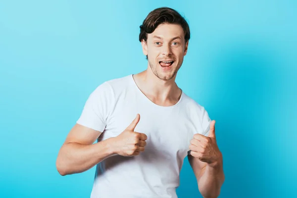 Mann im weißen T-Shirt zeigt Daumen hoch und blickt in Kamera auf blauem Hintergrund — Stockfoto