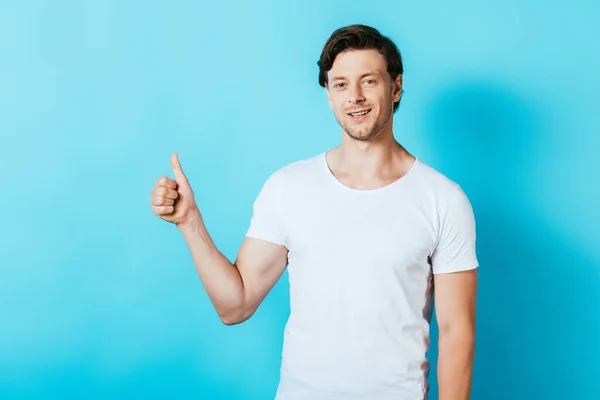 Молодий чоловік у білій футболці показує великий палець на синьому фоні — стокове фото