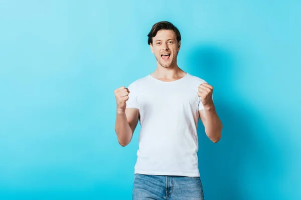 Homem de t-shirt branca olhando para a câmera e mostrando sim gesto no fundo azul — Fotografia de Stock