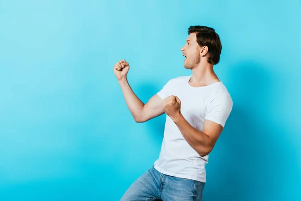 Homme excité en t-shirt blanc montrant geste ouais sur fond bleu — Photo de stock