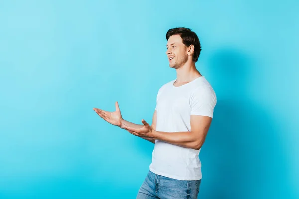 Young man in white t-shirt pointing with hands and looking away on blue background — Stock Photo