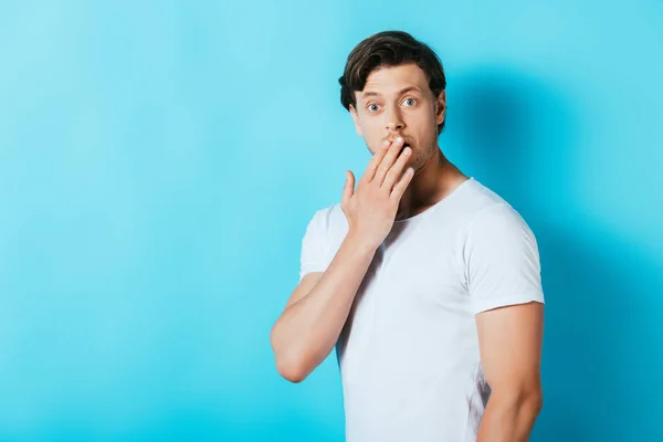 Schockierter Mann in weißem T-Shirt bedeckt Mund mit Hand auf blauem Hintergrund — Stockfoto