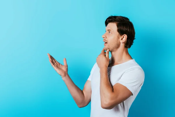 Hombre pensativo en camiseta blanca apuntando con la mano sobre fondo azul - foto de stock