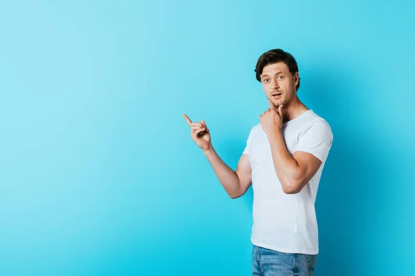 Homem pensativo em t-shirt branca apontando com o dedo no fundo azul — Fotografia de Stock