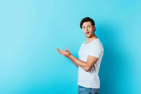 Homem animado em t-shirt branca apontando com as mãos no fundo azul com espaço de cópia — Fotografia de Stock