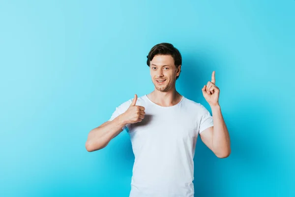 Jovem de t-shirt branca mostrando polegar para cima e tendo ideia sobre fundo azul — Fotografia de Stock