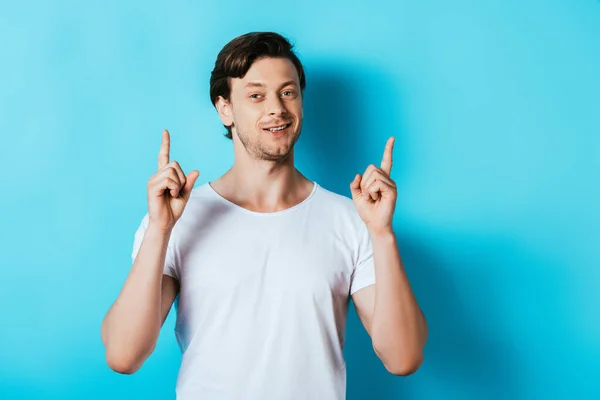 Hombre joven en camiseta blanca señalando con los dedos hacia arriba sobre fondo azul — Stock Photo