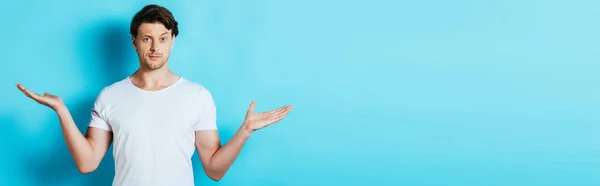 Cultivo horizontal de hombre confuso en camiseta blanca apuntando con las manos sobre fondo azul - foto de stock