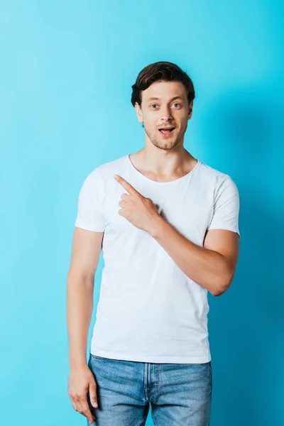 Homme excité en t-shirt blanc regardant la caméra tout en pointant du doigt sur fond bleu — Photo de stock