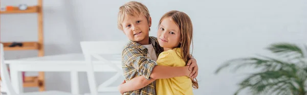 Hermano y hermana en ropa casual abrazando y mirando a la cámara mientras están de pie en casa - foto de stock