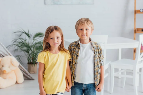 Chica en camiseta amarilla con hermano en camisa a cuadros mirando a la cámara mientras está de pie en casa - foto de stock