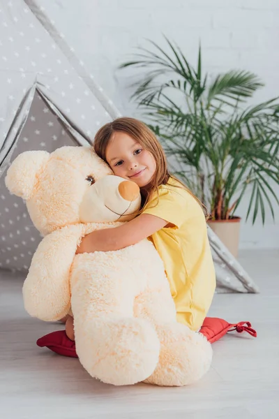 Niña en camiseta blanca abrazando oso de peluche mientras está sentado en el suelo cerca de la tienda de juego - foto de stock