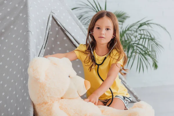 Criança em camiseta amarela olhando para a câmera enquanto joga médico com ursinho de pelúcia e estetoscópio perto de crianças wigwam — Fotografia de Stock